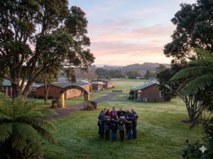 A wide-angle landscape shot of the Tuakau College campus entrance at dawn. A small group of teachers and community members are seen embracing and talking in a support circle on the school lawn. Soft, morning light filters through large NZ native trees (like Pōhutukawa). No recognizable faces.