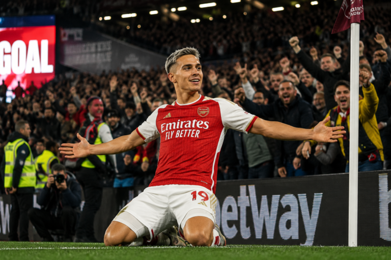 Leandro Trossard celebrating a late goal in front of jubilant away fans at London Stadium, Arsenal red kit, evening stadium lights, professional sports photography